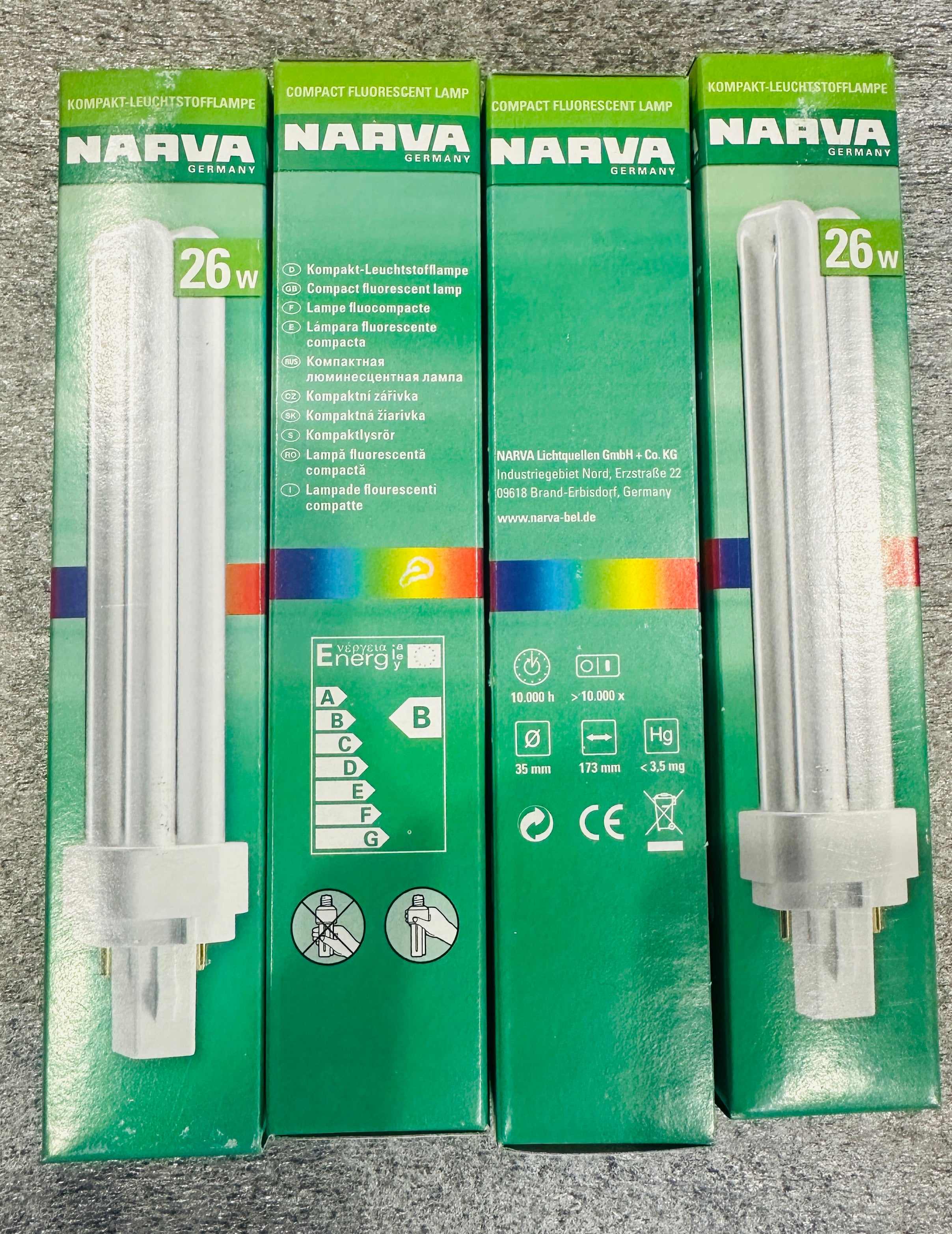 Three Narva 26W compact fluorescent light bulbs in their original green retail packaging, arranged side-by-side on a gray textured surface.